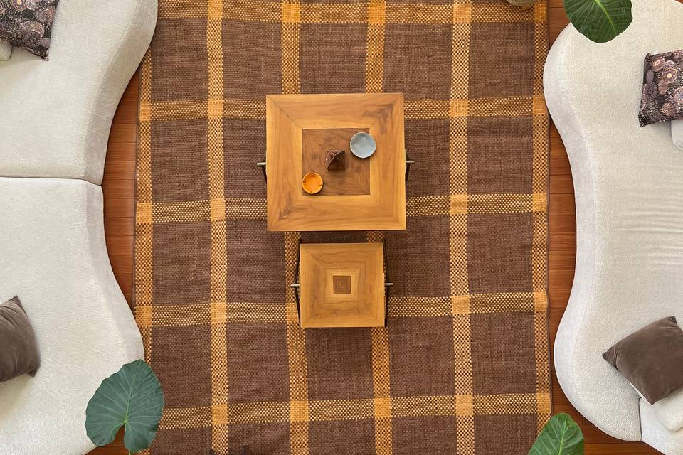 A top-down view of a brown and beige plaid area leather woven rug styled in a modern living room setting with curved white sofas, wooden coffee tables, potted plants, and a wooden floor. Main feature is the unique woven rug